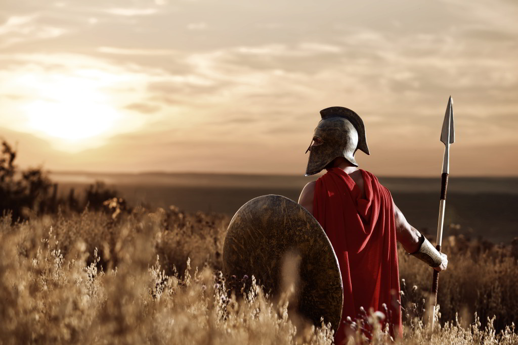 Ein Krieger steht mit einem Speer in der rechten Hand in einem Feld und beobachtet den Sonnenuntergang.