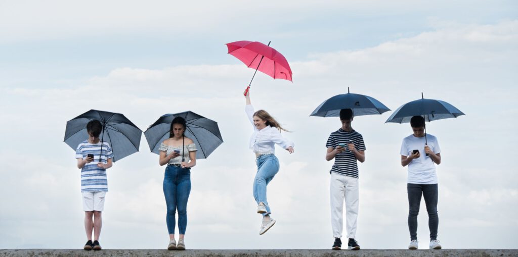 Bild von 5 Personen mit Regenschirmen die in einer Reihe stehen und in Mobiltelefone, blicken, eine Frau in der Mitte hat einen roten Regenschirm und springt in die Höhe.