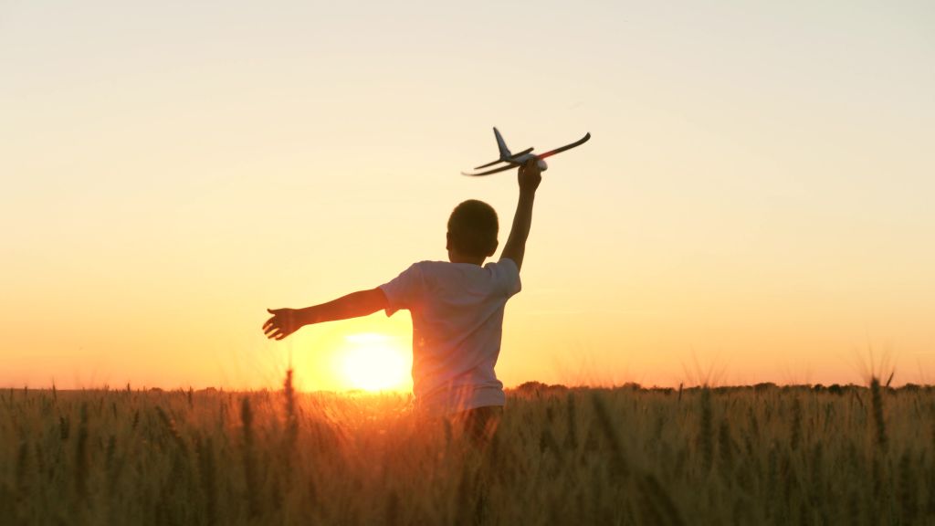 Ein Kind läuft mit einem Spielzeug-Flugzeug durch eine Feld Richtung Sonnenuntergang.