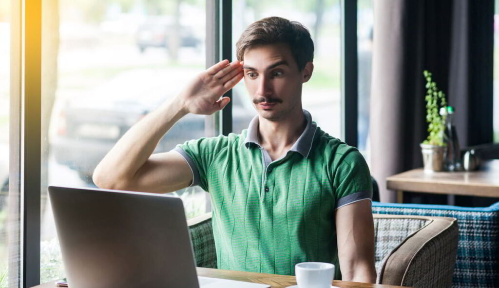 Mann in grünem Poloshirt salutiert vor einem Laptop in einem Café.